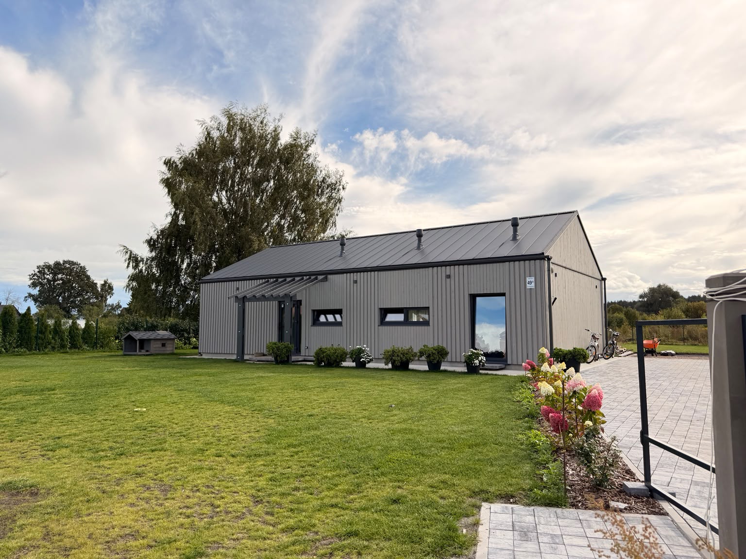 Completed grey timber home with a green lawn, paved terrace, pergola and flower beds